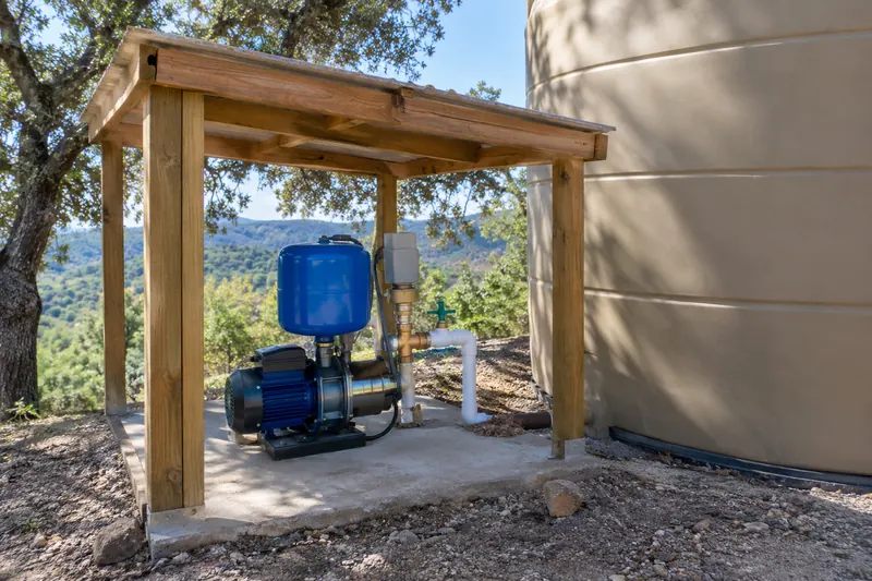 Small blue electric booster pump with a compact blue pressure tank, gray pressure switch and brass and PVC fittings on a concrete pad under a wooden lean-to beside a large tan water tank, wooded hills behind