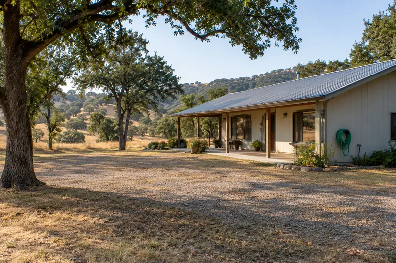 Single-storey ranch house with a metal roof and covered porch, surrounded by a wide cleared zone of short dry grass and gravel, oaks limbed up high, a green hose on a wall reel, tall golden grass and oak hills beyond
