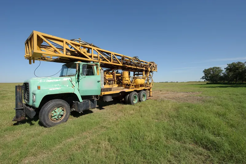 Older mint-green truck carrying a yellow folded drilling mast and rig machinery, parked in a grassy pasture under a clear blue sky