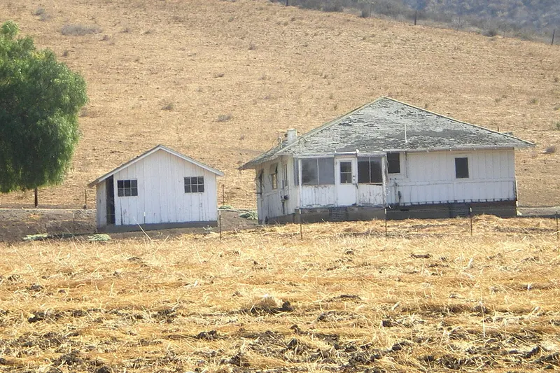 Weathered white board-and-batten ranch house with a hipped shingle roof and a small outbuilding, set against a dry golden hillside above a stubble field
