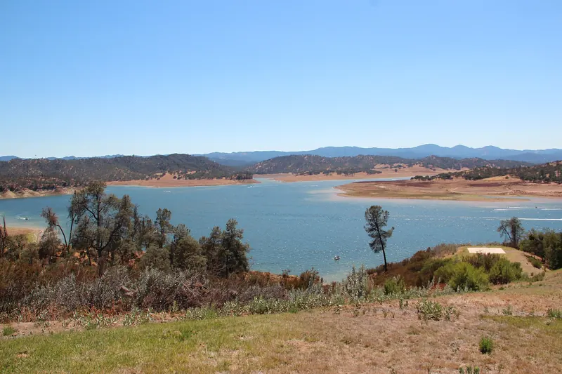 A lake well below its normal level, with wide bands of bare tan shoreline under oak- and pine-covered hills and dry brush in the foreground