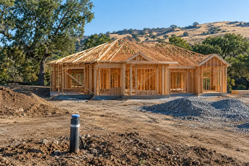 Freshly framed single-storey house with roof trusses on a graded dirt lot, a newly capped well casing in the foreground, gravel piles, oaks and golden hills behind