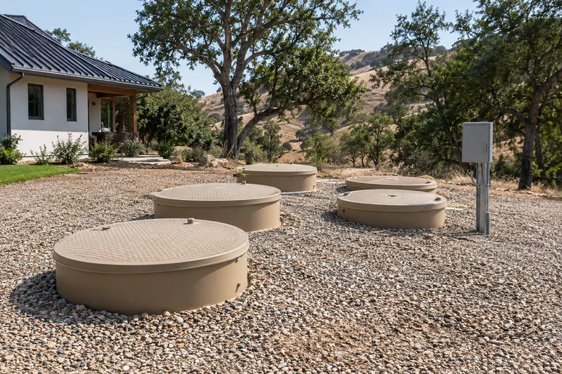 Several round tan plastic septic access risers with lids set in a gravel yard beside a newer white rural house, a gray control box on a post, oaks and dry hills behind
