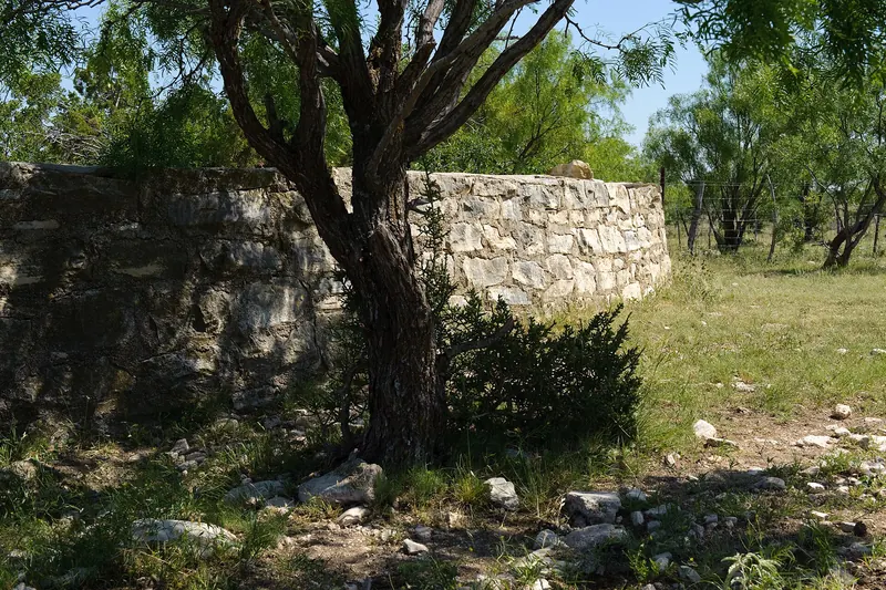 Curved stone-and-mortar wall of an old round ranch water storage tank shaded by a mesquite tree, with grass, rocks and a barbed-wire fence alongside