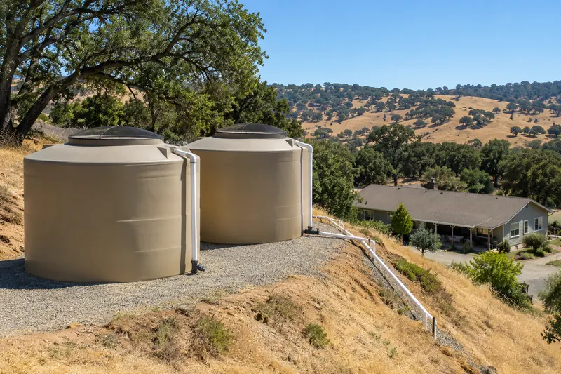 Two large tan polyethylene water tanks with screened domed lids on a gravel pad cut into a hillside, white PVC lines running down the slope toward a ranch house among oaks and golden hills