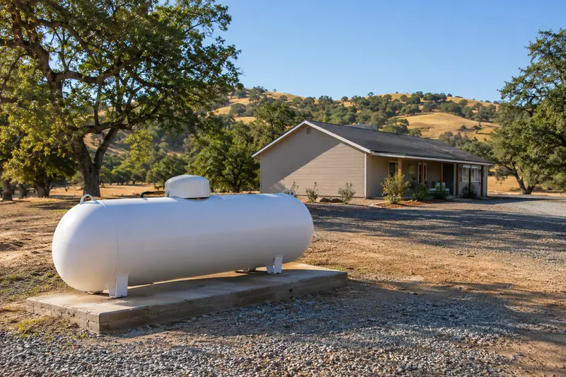 White horizontal propane tank on a concrete pad in a cleared gravel area, set well away from a single-storey rural house under oaks