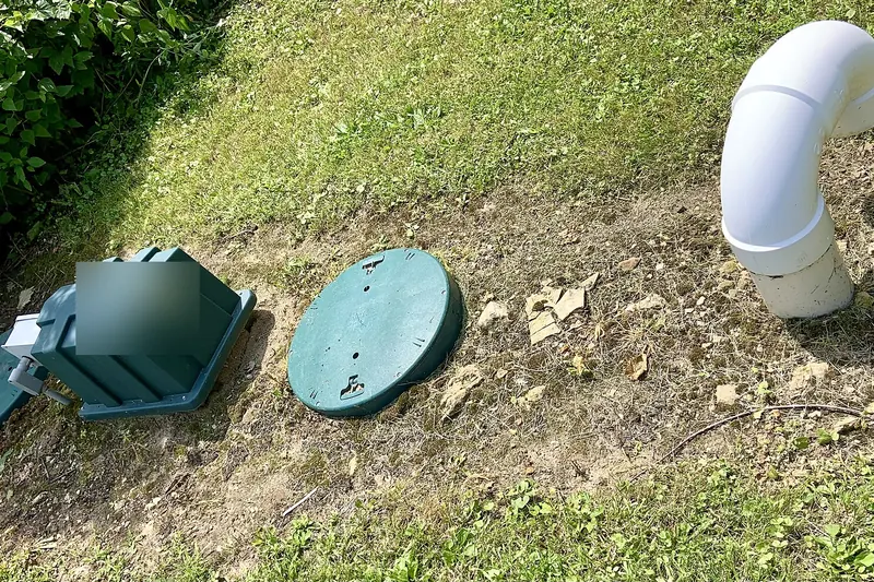 Green plastic lids and an equipment housing of a septic treatment unit set in a strip of bare soil in a lawn, with a white vent pipe elbow rising at the right
