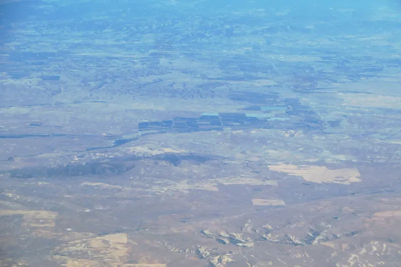 Hazy aerial view of dry tan hills and a block of irrigated farm fields along a valley, with more fields and ridges fading into the distance