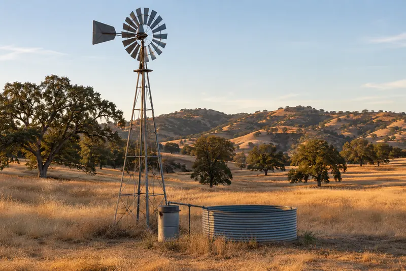 Old steel farm windmill over a capped well casing beside a round galvanized stock tank in a golden pasture with valley oaks